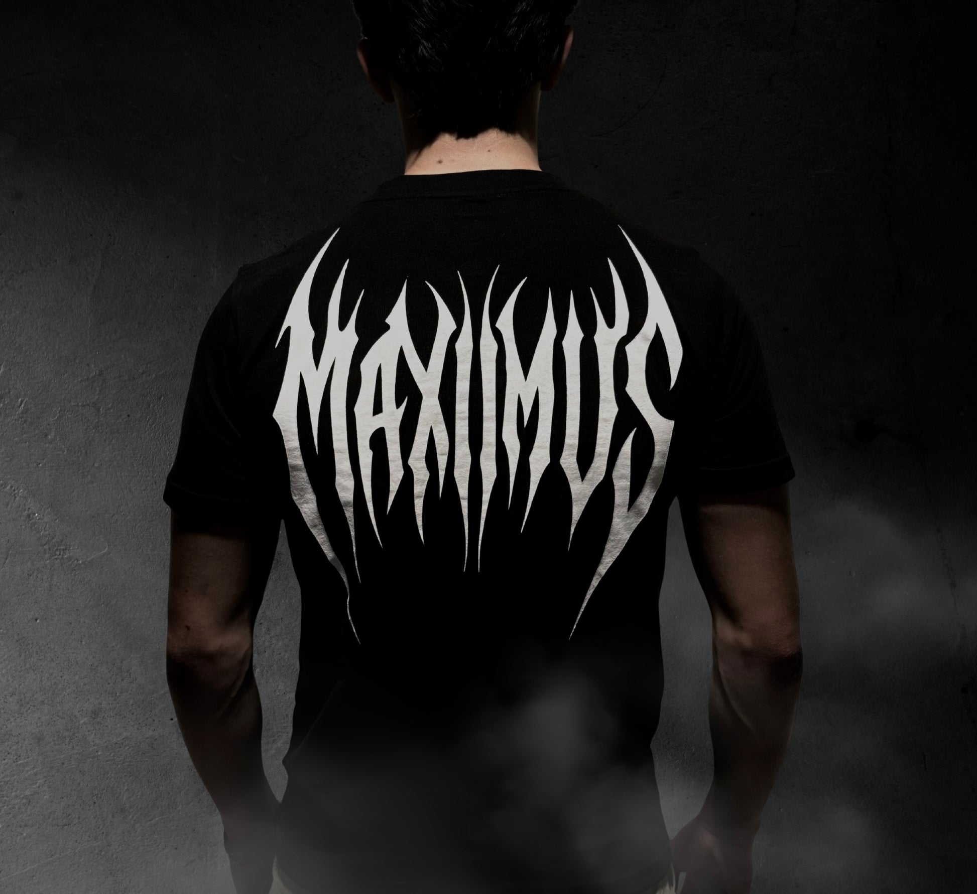 Person wearing a black t-shirt with 'MAXIMUS' design on the back against a dark background